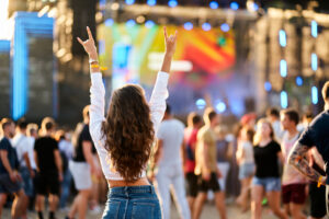 Back view of young woman with raised hands enjoys live music at sunny beach festival. Happy crowd around stage, summer event vibe. Fan cheers band, fun outdoor concert. Casual denim, festival fashion.