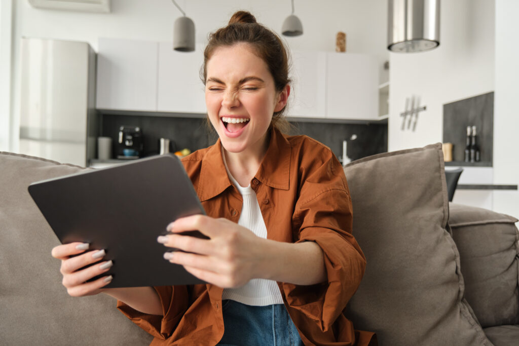 Portrait of excited young woman with digital tablet, sitting on couch, laughing and smiling, winning on video game, spending time at home
