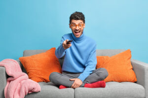 People leisure pastime concept. Overjoyed unshaven adult man sits in lotus poses on sofa holds remote control and watches funny show on television feels entertained enjoys lazy weekend at home