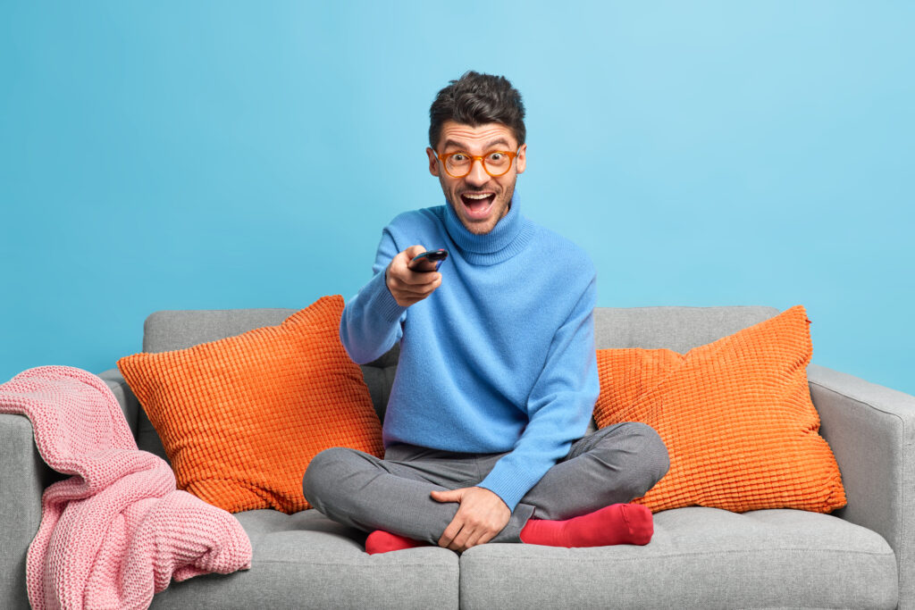 People leisure pastime concept. Overjoyed unshaven adult man sits in lotus poses on sofa holds remote control and watches funny show on television feels entertained enjoys lazy weekend at home